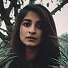 Portrait of a young woman with long dark hair and serious expression standing outdoors near greenery.